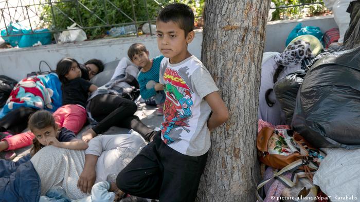 A family sits on a sidewalk in Athens, surrounded by bags