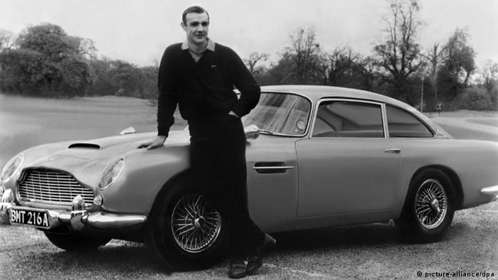 شين كونري في دور العميل السري 007، حوالي عام 1964 أمام سيارته من نوع أستون مارتن. 
