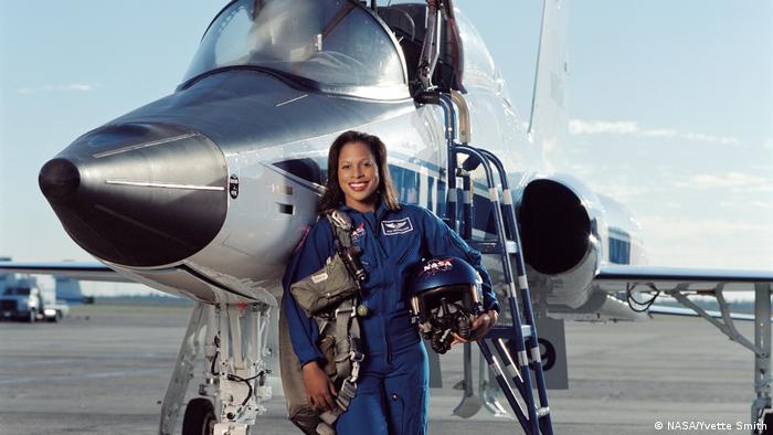 A woman leans against a plane wearing a blue NASA jumpsuit and cradling her helmet