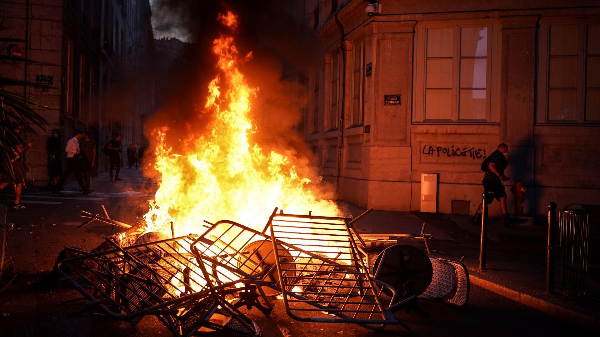 النار تشتعل في وسط طريق أغلقه المتظاهرون ضد عنف الشرطة في مدينة ليون الفرنسية وكُتب في الخلفية "الشرطة تَقتل" إثر مقتل الفتى ناهل بضواحي باريس، 30 يونيو/حزيران 2023.